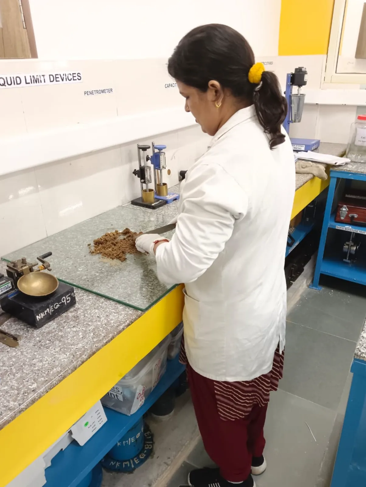 NKMPV soil testing technician preparing soil sample for Atterberg / Liquid Limit / Plastic Limit testing per IS 2720 Part 5 in the NABL-accredited soil-testing lab in Pinjore, Haryana - Casagrande device and penetrometer station visible on the bench.